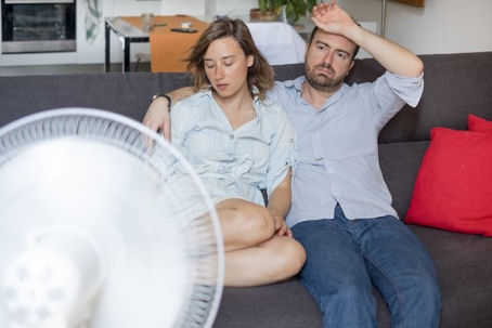 A man and a woman sit on a couch in front of a fan